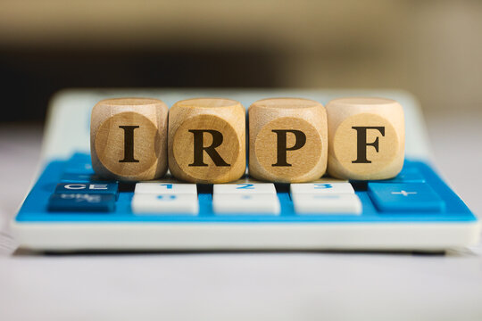 A sigla IRPF de Imposto sobre a Renda das Pessoas Físicas em Português do Brasil escrita em dados de madeira que estão sobre uma calculadora azul. Economia brasileira.