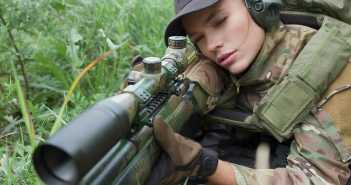 serious woman in cap, headset, tactical gear aiming with a sniper rifle sitting on the grass, training to shoot with gun, slow motion side view