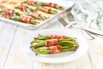 Homemade roasted bundles of green beans wrapped in bacon on a ceramic plate on a white wooden table, close up.