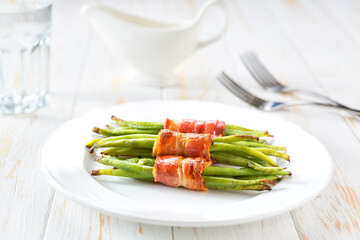 Baked  bundles of green beans wrapped in bacon in a ceramic plate on a white wooden table, close up.