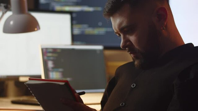 Programmer taking notes in the office. Selective focus of bearded man writing in the notebook at his workplace. Work routine, work in the office