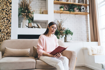 Positive ethnic woman sitting with book on sofa in living room