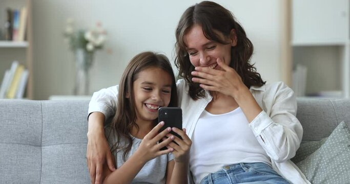 Cute preteen daughter have fun with mom sit on sofa, enjoy smartphone usage, having fun, using new mobile app, look at mobile phone screen watching videos on modern gadget, laugh, sit on sofa at home