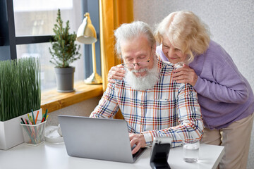 caucasian retired couple, 70s man and woman in living room browsing internet on modern laptop,...
