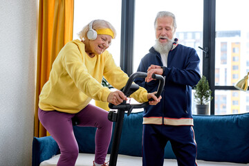 Fototapeta na wymiar Retired lady training on stationary bicycle at home, with personal trainer gray haired man. Senior adult doing workout and physical exercise while using cardio bike machine for wellness and endurance