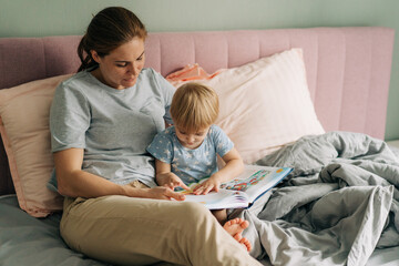 Mom reads a book with her little daughter lying in bed.