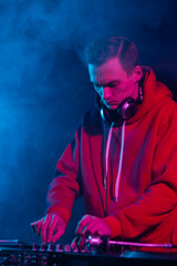 Cool young hip hop DJ in red hoodie mixing vinyl records on a party in night club. Professional disc jockey plays set in smoke on stage