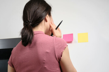 Oriental young woman holding pen and thinking about what to write on generic reminder note stuck onto fridge in house