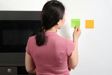 Oriental young woman sticking multicolored generic reminder notes onto fridge in house