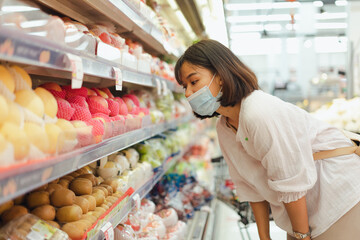 Portrait of healthy young Asian woman with casual clothes is wearing medical face mask when buy fresh organic fruit to make healthy food with new normal lifestyle  and social distancing at supermarket
