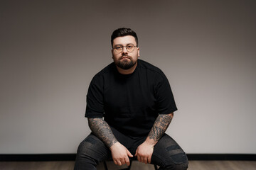 Content young plus size bearded male model in black outfit and eyeglasses with tattooed arms sitting on chair against gray background in studio