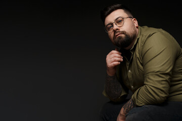 Thoughtful young plump tattooed male model in eyeglasses and green shirt looking at camera and touching beard against black background in studio
