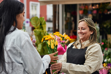 Young Latina woman purchasing vibrant flowers and plants from a street vendor's stall, handed by...