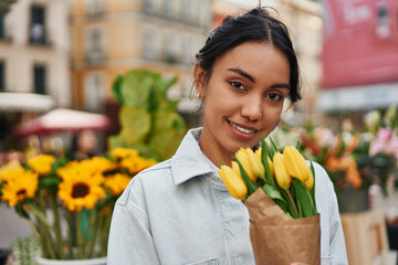 Young Latina woman smiling while buying yellow tulips from a street vendor's stall, adding warmth...