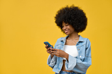 A hipster girl with afro haircut enjoy online communication and online services, wear trendy jeans jacket, scroll social media apps on cell phone device isolated with copy space on yellow background - obrazy, fototapety, plakaty