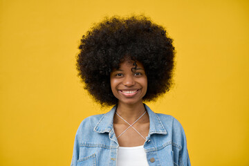 Obraz na płótnie Canvas A portrait headshot of a young happy smiling woman with a big afro hairstyle on her head on yellow orange background. A cheerful stylish hipster teenage female girl in jeans jacket looking at camera.
