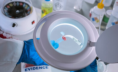 Police scientist analyzes bloody evidence in vial to discover suspect's DNA in a magnifying glass, concept image
