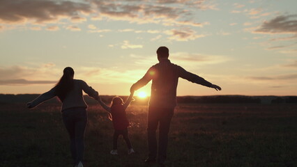 Fototapeta na wymiar Mom, dad and daughter are playing on field, child is jumping on green grass and holding hands of parents. Teamwork. Happy family runs in park holding hands in summer at sunset. Happy healthy childhood