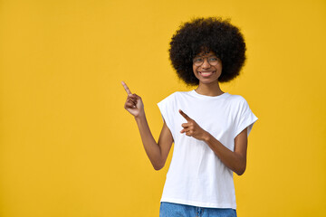 Portrait of teenage african american smart student girl in eyeglasses and with curly afro hairstyle stand and point on copy space for advertising, looking at camera and smiling on yellow background.