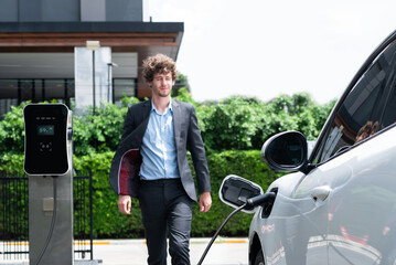 Progressive businessman wearing black suit with electric car recharging at public parking car...