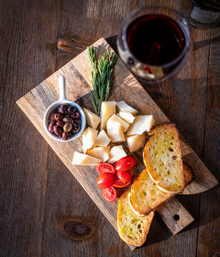 Flat lay food photograph of bruschetti with cheese, cherry tomatoes, olives and red wine over a wooden board on brown background