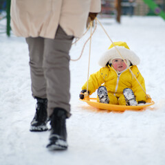 Toddler baby rides in the snow on icesled, a winter playground. Mother woman sledding baby boy in yellow snowsuit. Kid age one year eight months