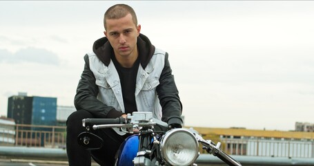 Handsome rider guy sitting on classic style cafe racer motorcycle at day time. Bike custom made in...