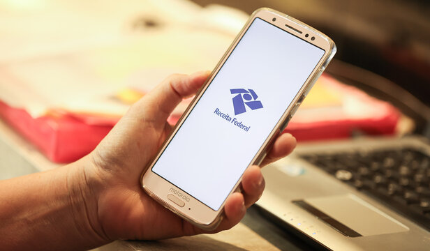 Uma pessoa segurando um telefone celular com a logo da Receita Federal do Brasil na tela. Imposto de renda, Governo Brasileiro.