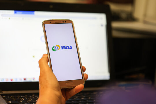 Uma mulher segurando um telefone celular com a lototipo do INSS na tela. Instituto Nacional do Seguro Social. Laptop ao fundo.	
