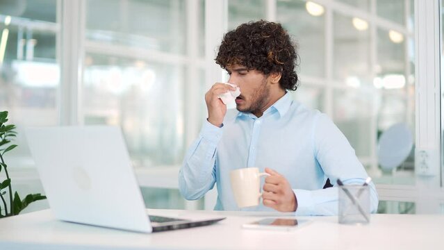 A sick young man has a runny nose working on a laptop while sitting at a workplace at a desk in a modern office. The worker wipes his nose with a handkerchief, drinks hot tea. You have a cold or flu