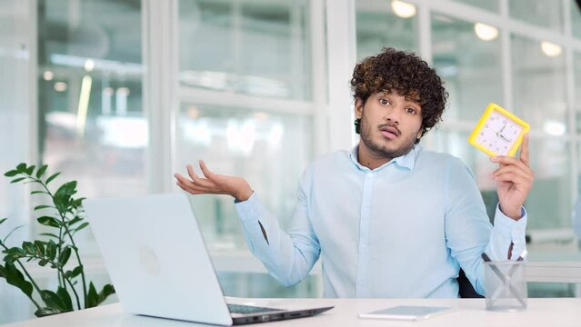 Young tired worker showing clock looking at camera while working on laptop while sitting at desk in modern office. Overworked man at work has no time and must finish the project before the deadline