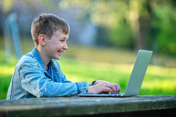 Cheerful teen boy studying online with a computer, outdoors. Boy on online lessons while outside....