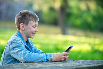 Young cheerful teen typing on mobile phone sitting behind a table in a park. Smiling child is texting on the phone in a garden, on a sunny summer day. Happy young boy playing on a cell phone outdoors.