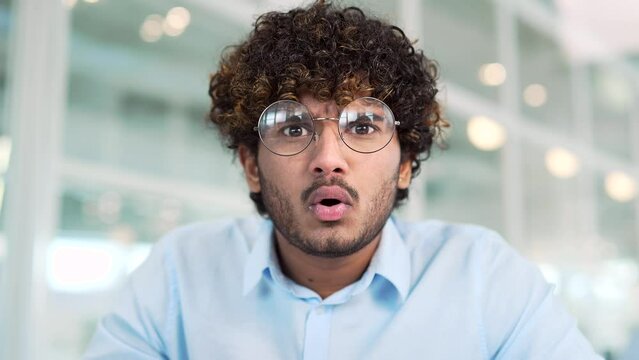 Webcam view. Close up of surprised puzzled young man in glasses looking at camera or laptop screen in office. The male spreads his hands, shrugs his shoulders, has a confused expression on his face