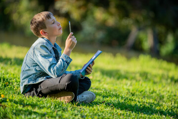 Thinking student with a textbook in his hands is sitting on a green lawn in a park. Pupil reading his notes and memorizing the text before a test. School boy writing in a notebook in a garden
