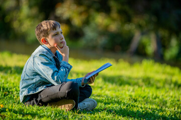 Student with a textbook in his hands is sitting on a green lawn in a park. Young boy reading his...