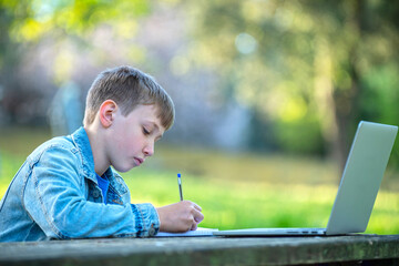 Boy is writing down notes outdoors. Scholar studying online with a computer. Boy on online lessons...