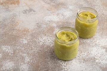 Baby puree with vegetable mix, broccoli, avocado in glass jar on brown concrete, side view, copy space