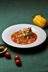 Shrimp with cherry tomatoes, avocado, cucumbers, and radishes in a red sauce on a white plate against a dark green background.