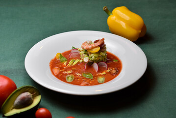 Shrimp with cherry tomatoes, avocado, cucumbers, and radishes in a red sauce on a white plate against a dark green background.