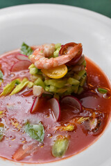 Shrimp with cherry tomatoes, avocado, cucumbers, and radishes in a red sauce on a white plate against a dark green background.