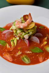 Shrimp with cherry tomatoes, avocado, cucumbers, and radishes in a red sauce on a white plate against a dark green background.