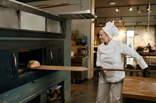 Young bakery worker using wooden shovel to take bread from stove oven