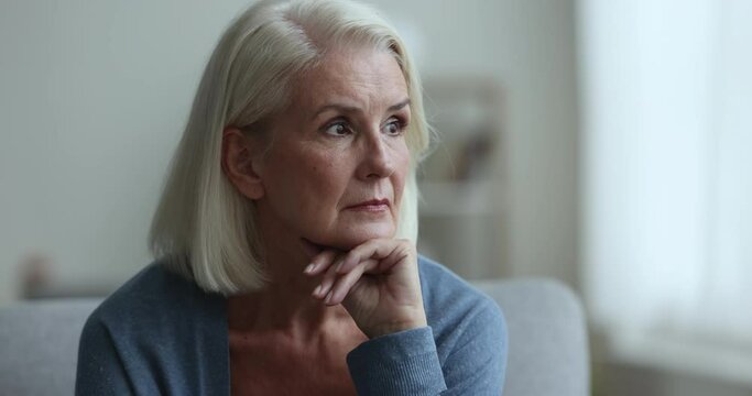 Close up sad, unhappy senior female sits on couch, looks in distance, have psychological problem, feels depressed, goes through older age crisis, worry about health, suffer from anxiety, miss of past
