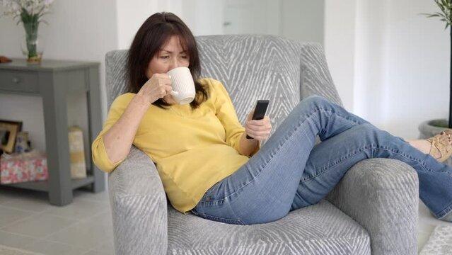 Attractive stylish woman enjoying drinking coffee using tv remote control changing television channels