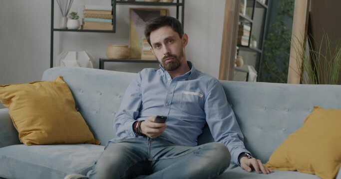 Portrait of attractive young man in trendy clothing watching TV using remote control relaxing on couch at home. People and entertainment concept.