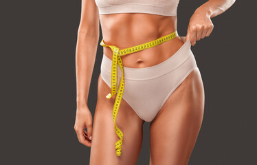 A slender dark-skinned girl in beige underwear measures her waist and hips with a centimeter on a dark background. The concept of weight loss and healthy lifestyle.