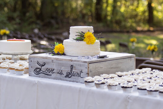 Backyard wedding with rustic country style cake stand holding wedding cake with yellow flowers on table with cupcakes, love is sweet in cursive.