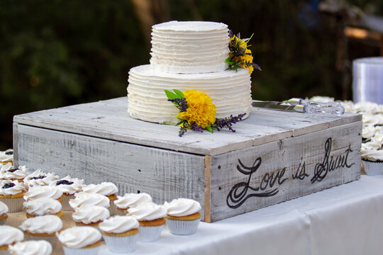 Love is sweet cursive rustic country style wedding cake with yellow flowers and cupcakes.
