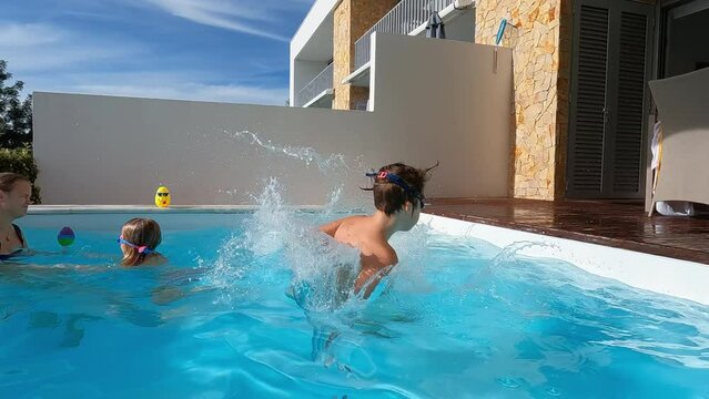 Slow-motion video of a boy in swimming goggles running and jumping into a pool at a private villa, his mother and sister dodge splashes, summer sun vacation